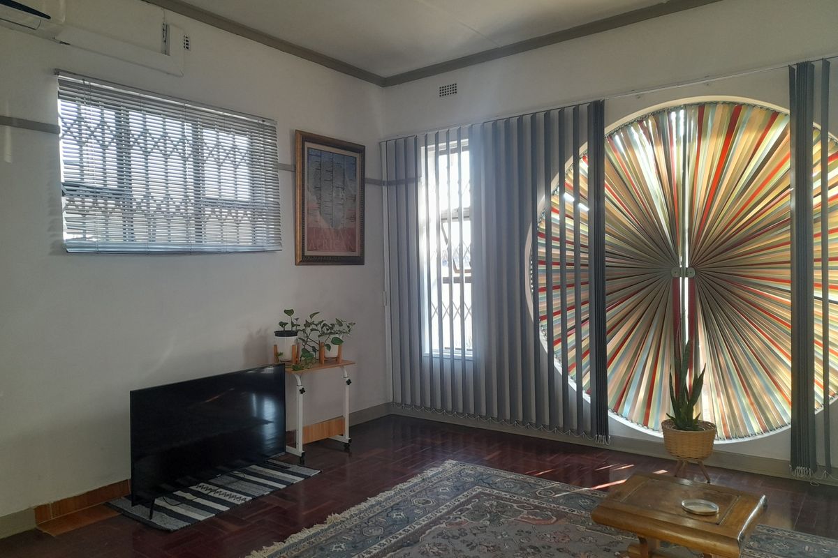 A bright living room featuring polished hardwood floors, a unique circular window with vertical blinds, and a wall-mounted air conditioning unit.