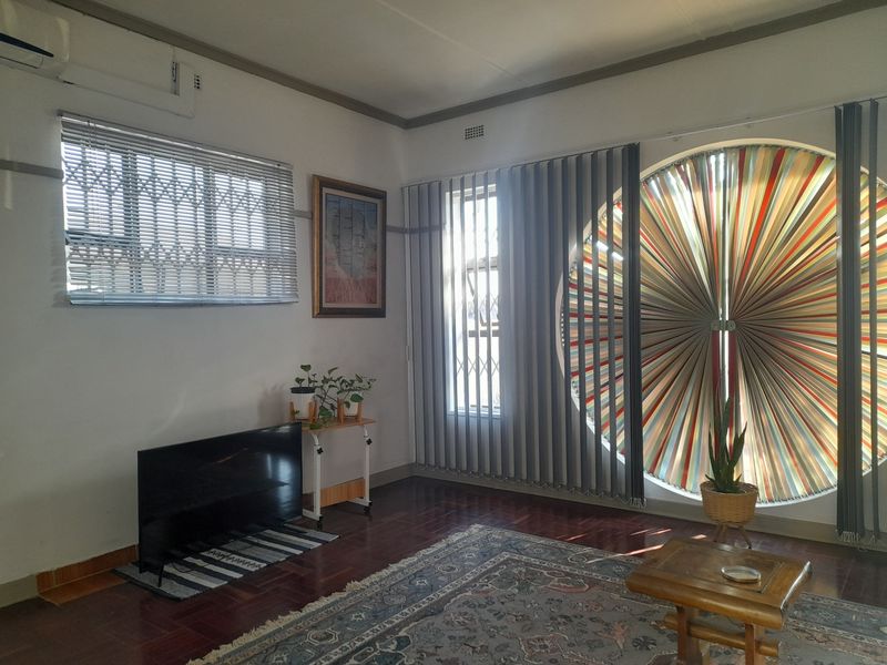 A bright living room featuring polished hardwood floors, a unique circular window with vertical blinds, and a wall-mounted air conditioning unit.