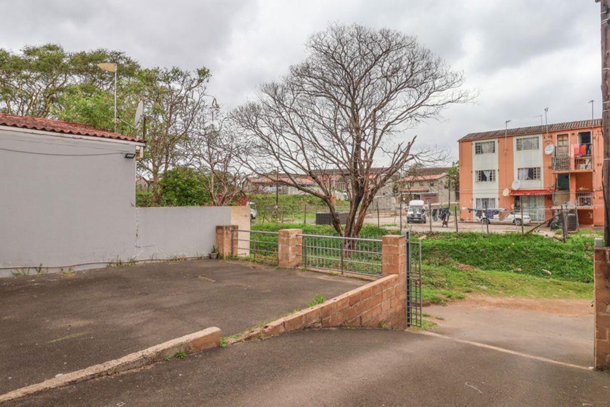 An exterior rear view showing an asphalt driveway, brick boundary wall, and metal gate, with surrounding residential buildings in the background.
