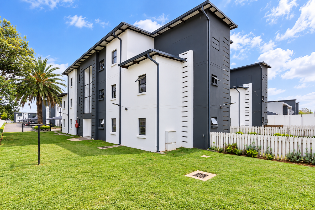 A well-maintained modern apartment building exterior featuring a clean white and dark grey facade, surrounded by a manicured lawn and white picket fencing.