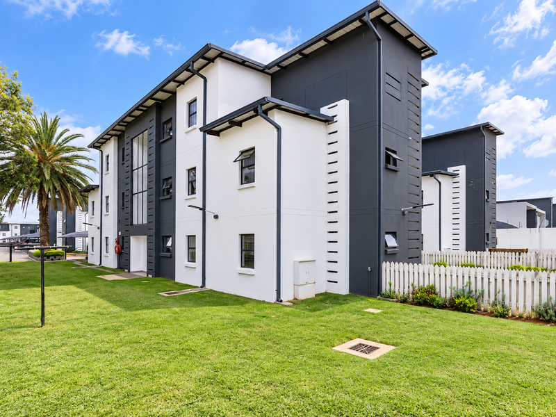 A well-maintained modern apartment building exterior featuring a clean white and dark grey facade, surrounded by a manicured lawn and white picket fencing.
