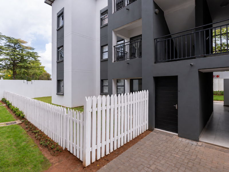 Modern apartment exterior featuring a private garden area with a white picket fence, paved entrance walkway, and contemporary dark grey and white facade.