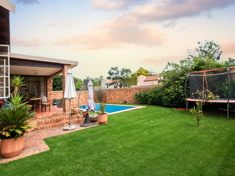 Rear garden featuring a covered brick patio, a swimming pool, and a well-maintained lawn enclosed by a brick boundary wall.