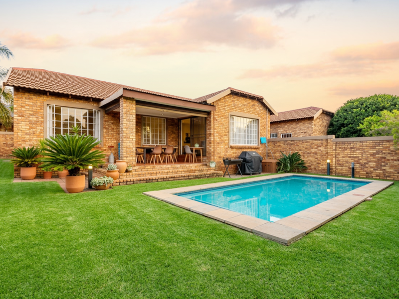A well-maintained rear exterior view of a brick townhouse featuring a private swimming pool, landscaped lawn, and a covered patio area.