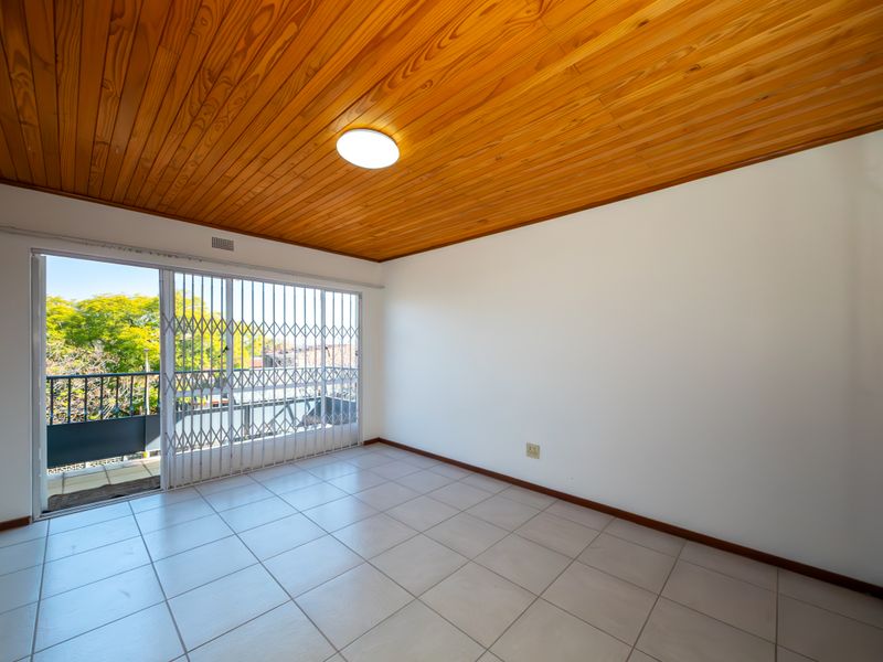 An empty living room featuring tiled flooring, a timber-paneled ceiling, and a sliding glass door leading to a balcony with security gate access.