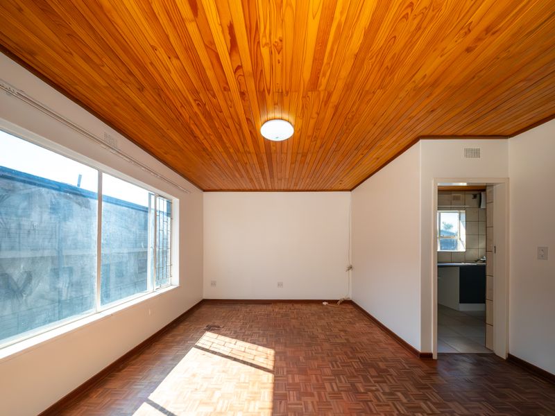 An empty living room featuring classic parquet flooring and a warm wooden tongue and groove ceiling, with natural light and access to the kitchen.
