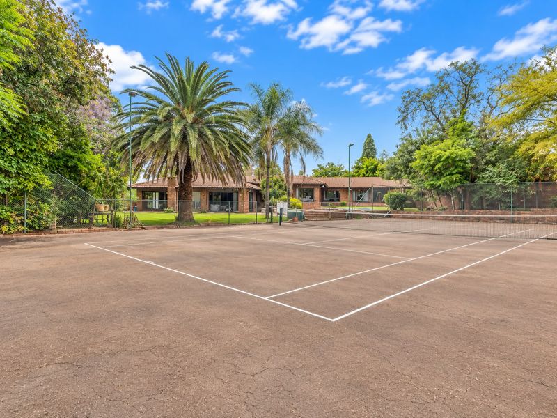 Private outdoor tennis court with chain link fencing and floodlights, set within a landscaped garden area.