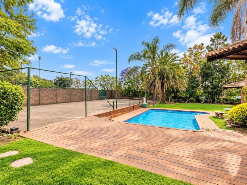 Outdoor recreational area featuring a private swimming pool, paved patio, and a fenced tennis court set within a landscaped garden.