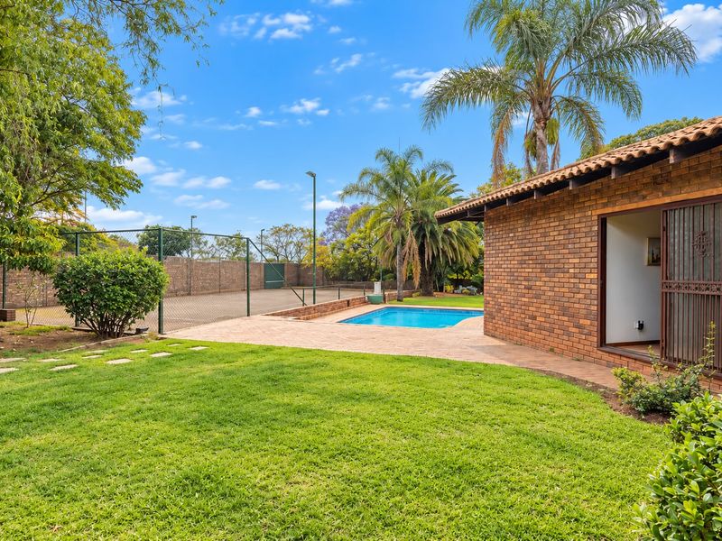 Rear exterior view featuring a brick house, swimming pool, and a private tennis court set within a landscaped garden.