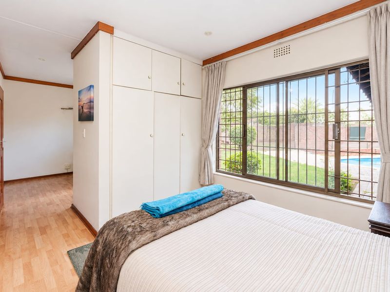 A bedroom featuring built-in cupboards, laminate flooring, and a large window with security bars overlooking the garden and pool area.