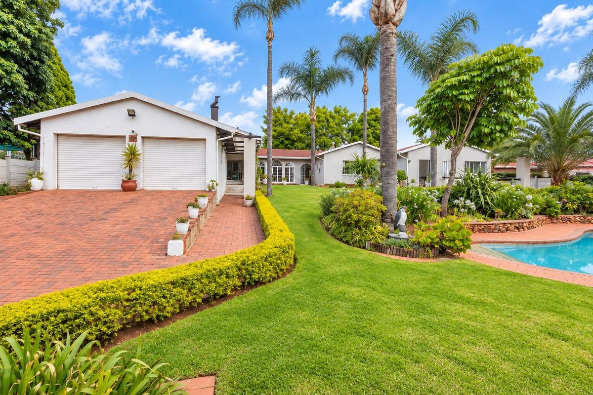Spacious rear exterior featuring a well-maintained garden, swimming pool, paved driveway, and a double garage with roller shutter doors.
