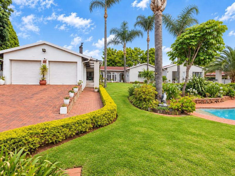 Spacious rear exterior featuring a well-maintained garden, swimming pool, paved driveway, and a double garage with roller shutter doors.