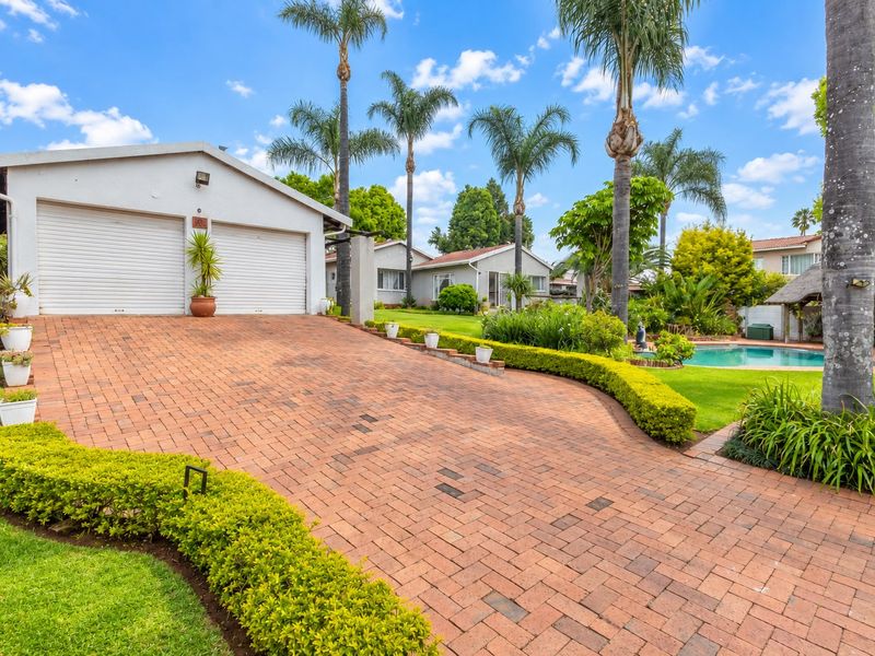 Front view of the property featuring a double garage with roller doors, a brick-paved driveway, and a well-maintained garden with a swimming pool.