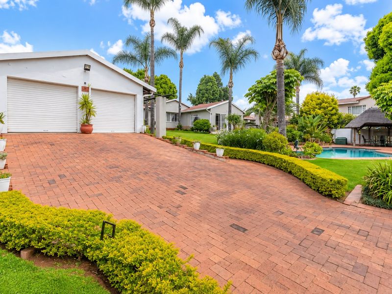 A well-maintained front exterior featuring a double garage, brick-paved driveway, landscaped garden, and a swimming pool with a thatched lapa area.