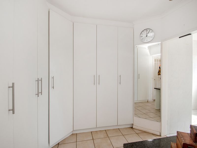 A clean bedroom space featuring extensive white built-in cupboards, tiled flooring, and a full-length mirror, offering ample storage and a bright, neutral aesthetic.