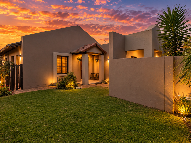 A well-maintained modern home exterior featuring a covered entrance, paved walkway, and landscaped garden, illuminated by warm evening lighting against a sunset sky.