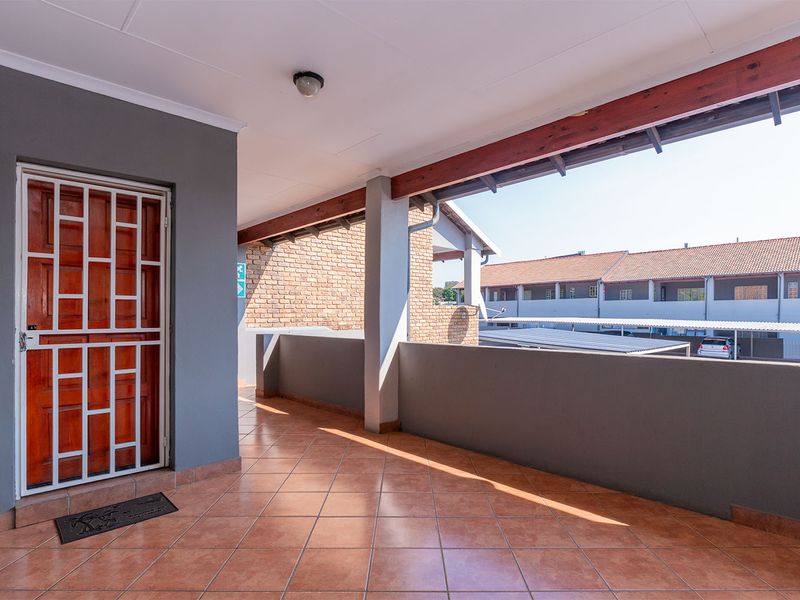 A covered balcony area featuring tiled flooring, a timber beam detail, and a secure entrance door, overlooking the apartment complex grounds.