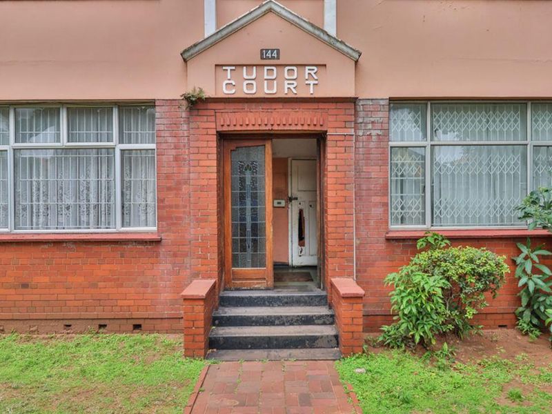 Front entrance of Tudor Court apartment building featuring classic brick facade, security gates, and paved walkway access.