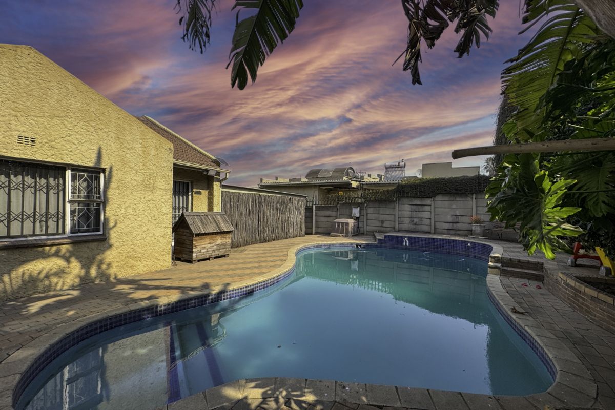Outdoor pool area featuring a paved patio, swimming pool, and secure boundary wall, set against a sunset sky.