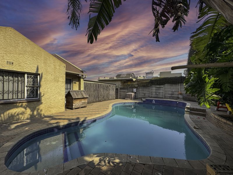 Outdoor pool area featuring a paved patio, swimming pool, and secure boundary wall, set against a sunset sky.
