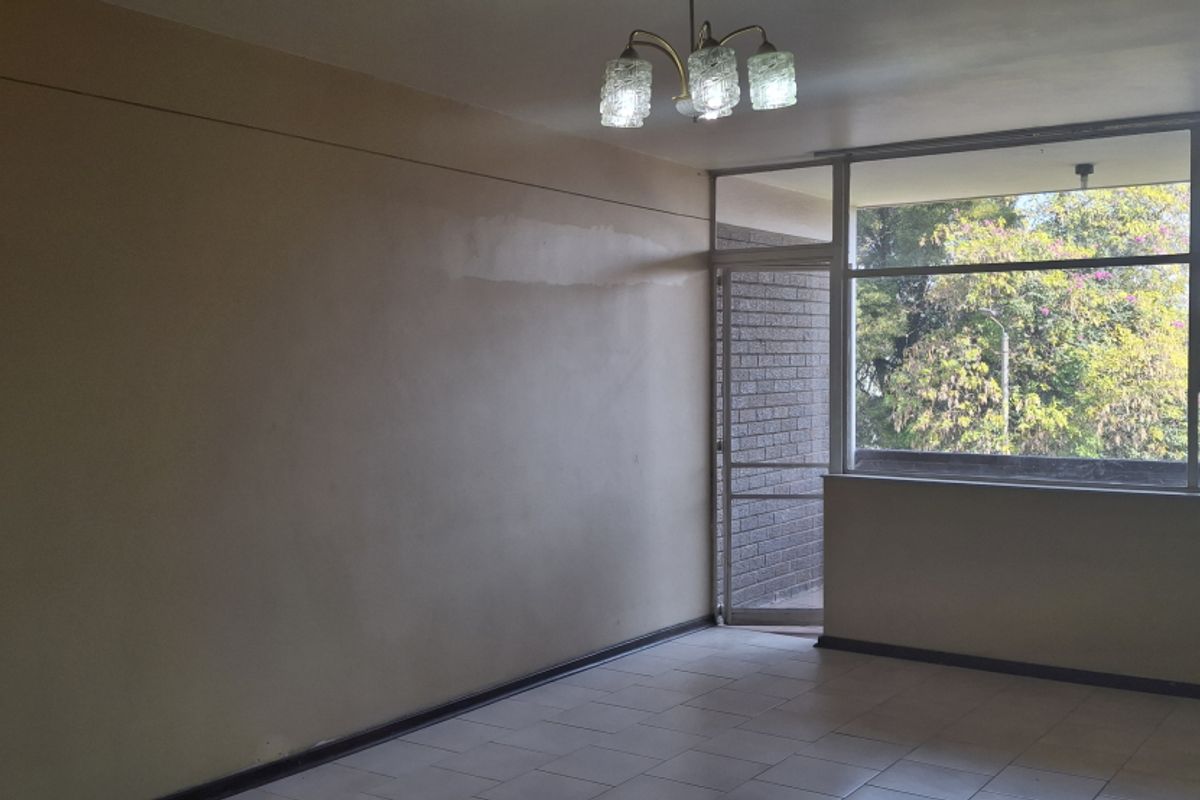 An empty living room featuring tiled flooring, a large window with natural light, and a brick feature wall, ready for renovation or staging.