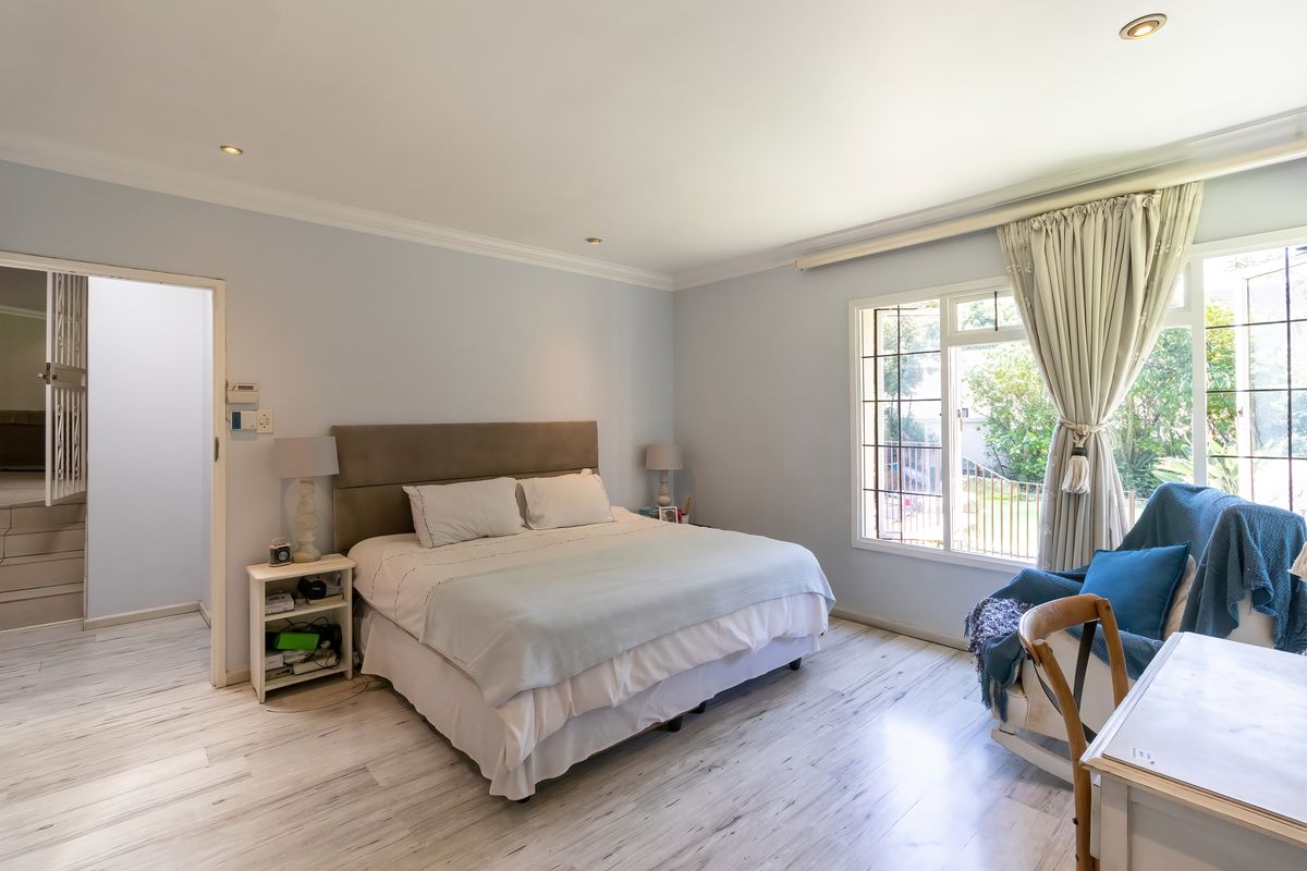 A well-lit bedroom featuring light-toned laminate flooring, recessed ceiling lighting, and a large window providing natural light and garden views.