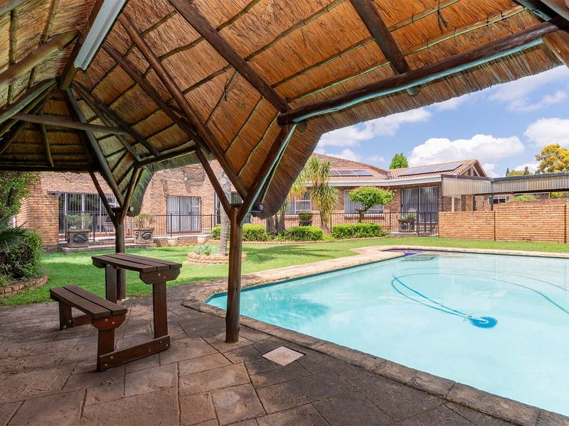 Outdoor entertainment area featuring a swimming pool and a traditional thatched lapa set within a private garden with brick boundary walls.