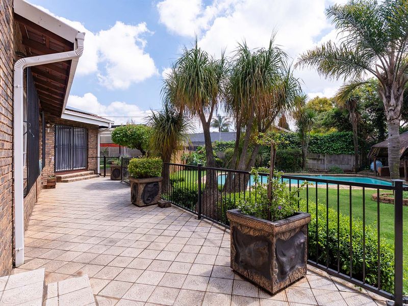 A spacious tiled patio overlooking a well-maintained garden, swimming pool, and a traditional thatched lapa, perfect for outdoor entertaining.