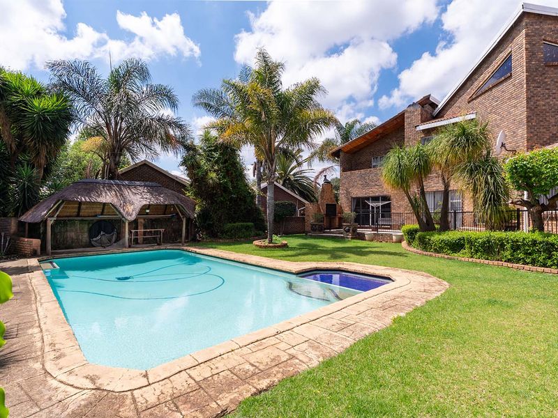 Spacious backyard featuring a swimming pool, a traditional thatched lapa, and a well-maintained garden set against a multi-story brick house.
