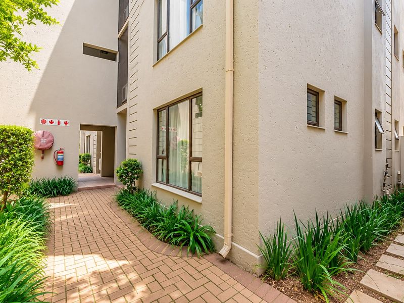 A well-maintained exterior rear walkway featuring paved paths, landscaped garden beds, and functional fire safety equipment against a modern apartment building facade.