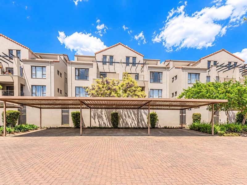 A well-maintained apartment complex exterior featuring a paved driveway and a modern covered parking structure with landscaped garden beds.