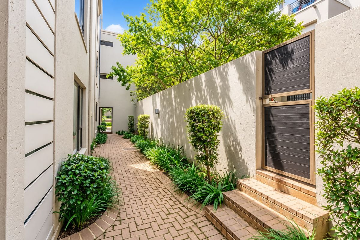 A well-maintained paved side walkway featuring a secure pedestrian gate, brick steps, and landscaped garden borders along the apartment building exterior.