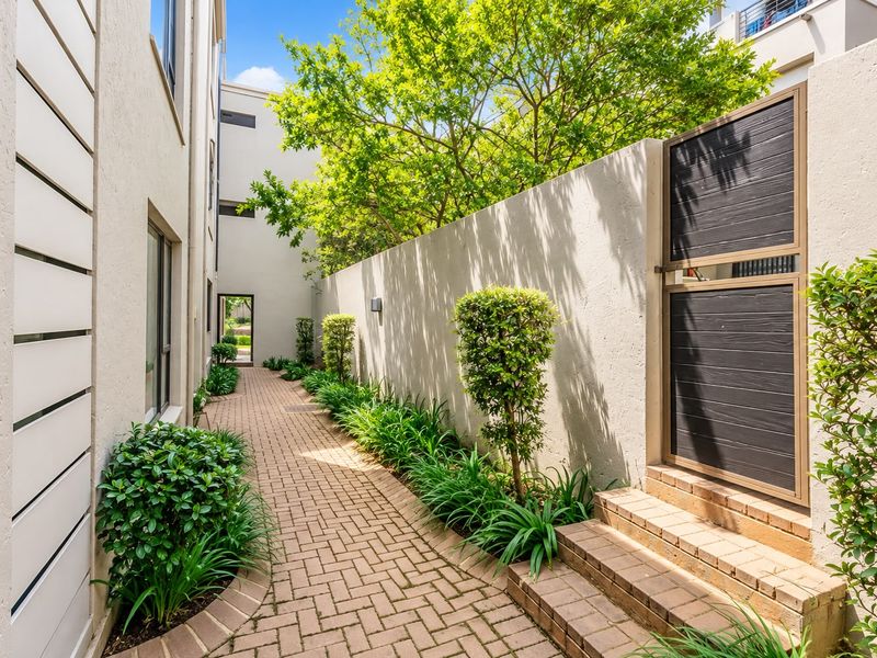 A well-maintained paved side walkway featuring a secure pedestrian gate, brick steps, and landscaped garden borders along the apartment building exterior.