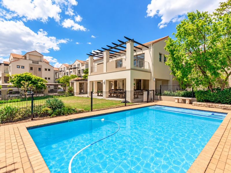 A well-maintained swimming pool area featuring a paved deck, perimeter fencing, and a covered patio with a pergola in a residential apartment complex.