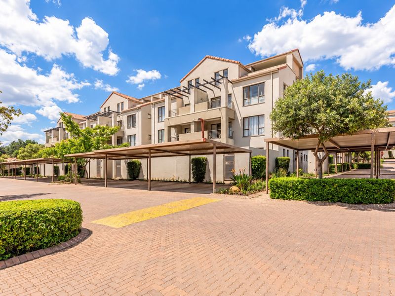 A well-maintained apartment complex exterior featuring paved driveway access, covered parking bays, and landscaped garden areas under a clear blue sky.