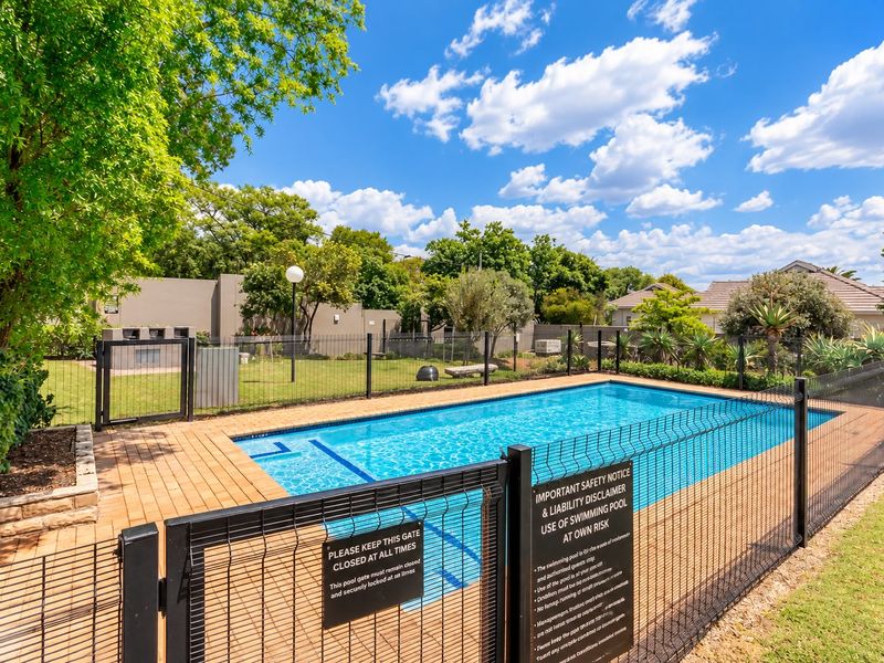 A well-maintained outdoor swimming pool area featuring a paved deck, safety fencing, and a built-in braai facility in the garden.