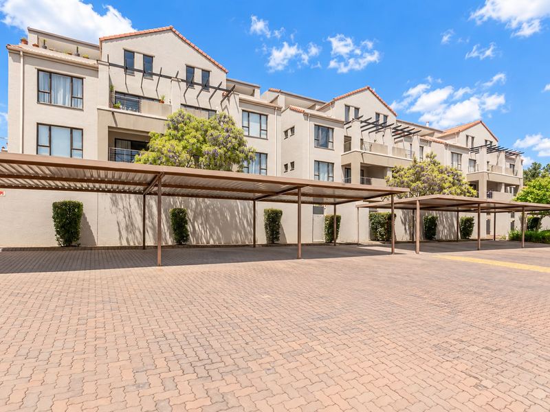 Exterior view of an apartment complex featuring covered parking bays, paved driveway, and multi-level residential units with balconies and pergola structures.