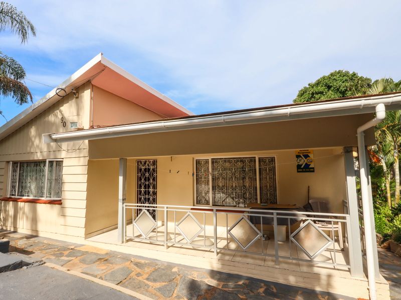 Front elevation of a single-story house featuring a covered patio, security gates, and window bars, set against a paved driveway and landscaped garden area.