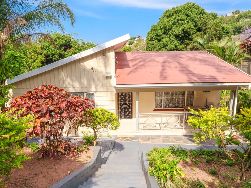 Front view of a charming house featuring a pitched roof, covered patio, and a concrete staircase leading to the entrance, surrounded by established garden greenery.