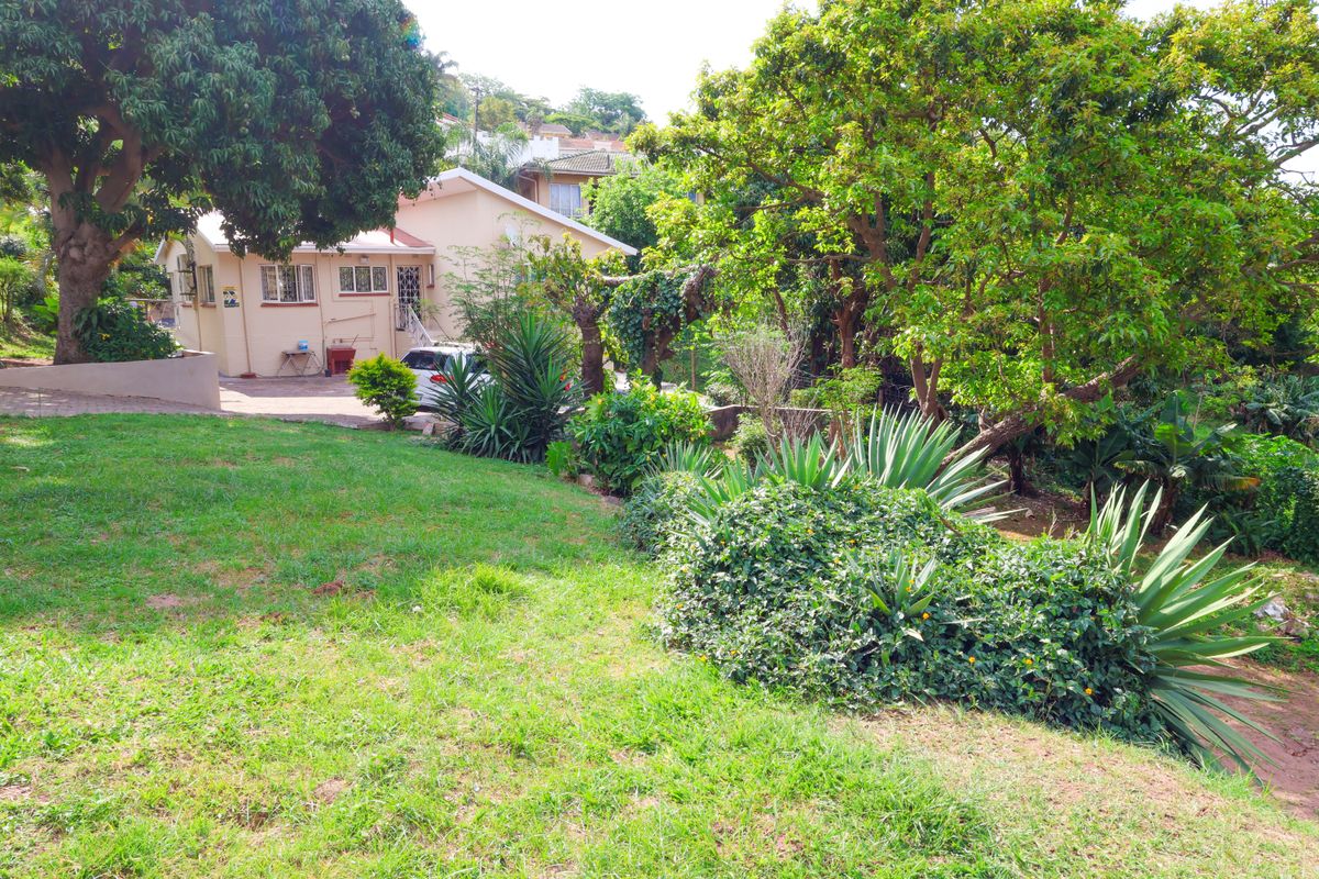 A view of the rear garden and property exterior, featuring mature trees, established landscaping, and a paved driveway area.