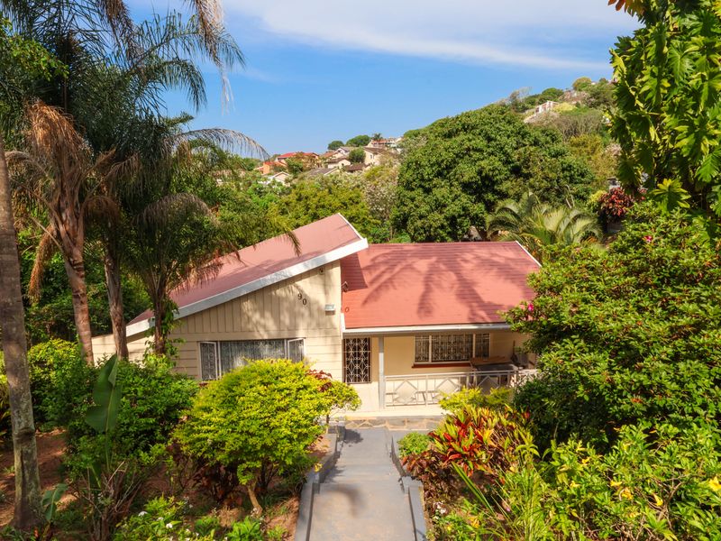 A charming residential property featuring a pitched metal roof and timber cladding, set within a lush, landscaped garden with a concrete pathway entrance.