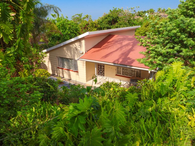 A view of the front elevation of a house featuring a pitched roof and a covered porch, surrounded by lush garden foliage.