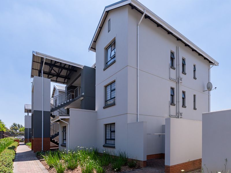 A well-maintained multi-story apartment building exterior featuring modern grey and white facade, brick detailing, and paved walkways with landscaped garden borders.