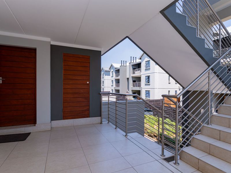 A clean and modern apartment entrance landing featuring tiled flooring, a stainless steel balustrade, and a staircase leading to upper levels.