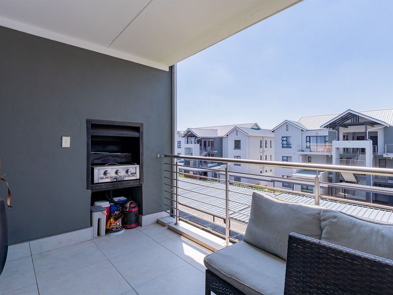 A modern covered balcony featuring a built-in braai, tiled flooring, and a stainless steel balustrade, offering an elevated view of the surrounding apartment complex.