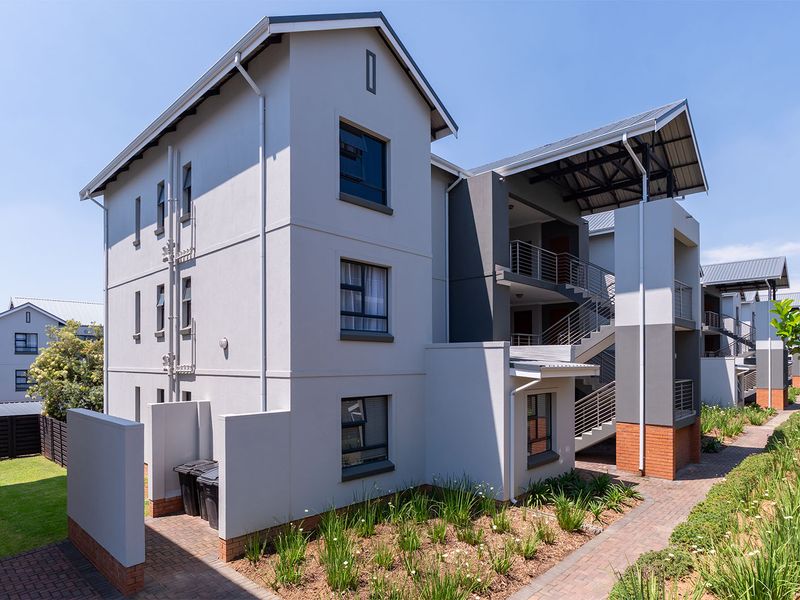 A modern multi-story apartment building exterior featuring a staircase, balconies with metal railings, and well-maintained landscaped garden areas with paved walkways.