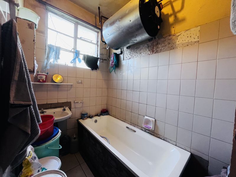 A dated bathroom featuring a bathtub, wall-mounted basin, and exposed electric water heater, requiring significant renovation and cosmetic updates to improve functionality and aesthetics.