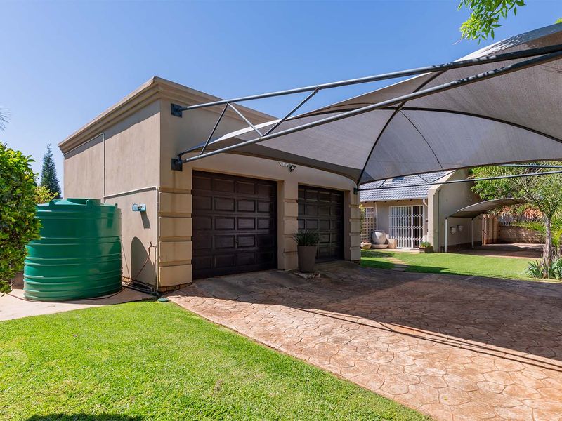 A well-maintained double garage featuring a shade cloth carport, paved driveway, and an integrated water storage tank set within a neat garden.