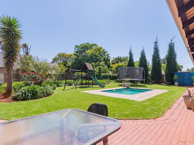 A well-maintained backyard featuring a swimming pool, lush lawn, landscaped garden beds, and a spacious brick-paved patio area under a covered veranda.