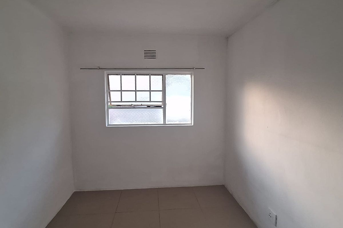 An empty, neutrally painted bedroom featuring tiled flooring, a window with security bars, and a central ceiling light fixture.
